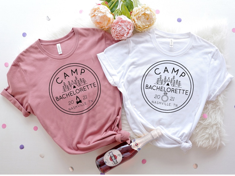 May include: Two white t-shirts with a black circle design that says "Camp Bachelorette 2021 Nashville, TN". One shirt is pink and the other is white. The design on the white shirt includes a ring icon.