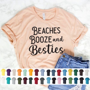 May include: A peach colored t-shirt with the text "Beaches Booze and Besties" in black. The shirt is shown on a white surface with a pair of blue jeans and a variety of t-shirt colors in the bottom of the image.