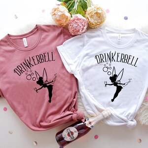 May include: Two t-shirts with the text "DRINKERBELL" and a silhouette of a fairy holding a glass of wine and a bottle of champagne. The shirts are in pink and white.
