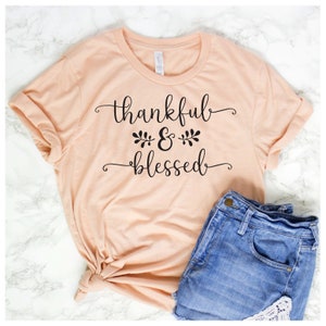 May include: A peach-colored t-shirt with the text "thankful & blessed" in black script font. The shirt is styled with a knot in the front and is paired with blue denim shorts.