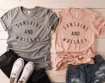 May include: Two t-shirts with the text "Sunshine and Whiskey" printed on them. One shirt is gray and the other is peach.
