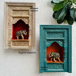 Peut inclure: Deux étagères murales décoratives en bois, une beige et une turquoise, chacune avec un motif sculpté. À l'intérieur de chaque étagère se trouve une petite statue d'éléphant dorée. Les étagères sont montées sur un mur blanc.