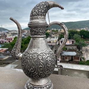 Puede incluir: Una tetera de plata con intrincados diseños florales y geométricos. La tetera tiene un pico largo y un asa curva. La tetera está decorada con una cadena y una tapa decorativa.