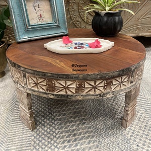 May include: A round wooden table with carved details and white painted accents. The table has four legs and a white marble top. A small plant in a black pot and a white tray with pink flowers are on the table. The table is on a gray and white patterned rug. The text "@Jeypore Souvenirs" is on the table.