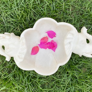 Puede incluir: Plato de servir de mármol blanco con forma de dos elefantes que se miran. El plato tiene un cuenco poco profundo en el centro y pétalos de flores rosas en su interior.