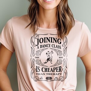 May include: A pink t-shirt with a black graphic design that reads "Joining a Dance Class is Cheaper Than Therapy". The graphic is styled to look like a vintage liquor label with a dancing couple silhouette.