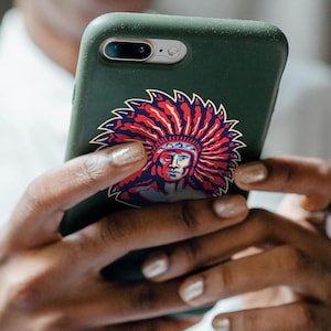 May include: A green phone case with a colorful sticker of a Native American man wearing a headdress. The sticker is in the center of the case.
