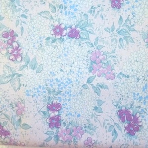 Op de afbeelding: Een bloemenpatroon met kleine witte en blauwe bloemen, en grotere paarse en roze bloesems. De bladeren zijn een gedempte zeegroene kleur, tegen een lichte achtergrond. Geschikt voor stof of behang.