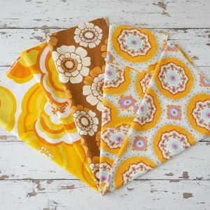 Op de afbeelding: Vier gevouwen vintage stoffen servetten met bloempatronen in tinten geel, oranje en bruin. De ontwerpen omvatten grote bloemen en geometrische vormen. De servetten zijn gerangschikt op een wit, verweerd houten oppervlak.