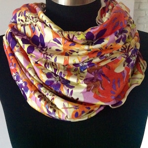Könnte beinhalten: Ein kreisförmiger Infinity-Schal mit einem lebendigen, mehrfarbigen Blumen- und Blattmuster. Der Stoff zeigt eine Palette aus leuchtendem Orange, tiefem Violett, zartem Rosa und blassem Gelb, angeordnet in einem dichten, überlappenden botanischen Design.