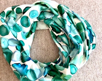 May include: An infinity scarf featuring a white background with a watercolor-style pattern of overlapping circular shapes in various shades of emerald green, forest green, teal, and light blue, creating a fluid and layered visual effect.