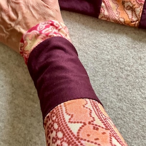 Könnte beinhalten: Ein langärmeliges Kleidungsstück mit abwechselnden Abschnitten aus einfarbigem, tiefem Burgunderrot und einem gemusterten Textil mit wiederkehrendem geometrischem Design in Orange-, Rosa- und Cremetönen. Die Ärmelkonstruktion zeigt deutliche Farbblöcke.