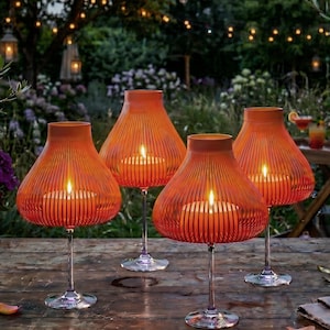 May include: Four orange candle holders with a ribbed design, each holding a lit candle. The candle holders are on clear stems and sit on a wooden table. The background features a garden setting with string lights.