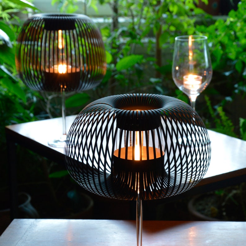 以下が含まれることがあります： 円形のワイヤーケージデザインの黒い金属製のテーブルランプが2つ。ランプは暖色系の黄色い光で点灯しています。ランプは木製のテーブルの上に置かれています。