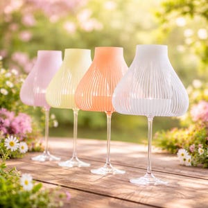 Könnte beinhalten: Vier Kerzenhalter aus Glas in Form umgedrehter Weingläser mit gerippten, glockenförmigen Schirmen in Pastellrosa, Gelb, Orange und Weiß. Jeder Halter hat einen schlanken Stiel und einen runden Fuß, konzipiert für kleine Teelichter.