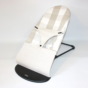 Puede incluir: Un asiento mecedor para beb&eacute;s a rayas beige y blancas con una base negra. El asiento tiene un asiento acolchado y un arn&eacute;s.
