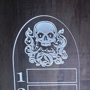 Może przedstawiać: Biała czaszka z białym ośmiornicą owiniętą wok&oacute;ł niej. Czaszka i ośmiornica znajdują się na czarnym tle.