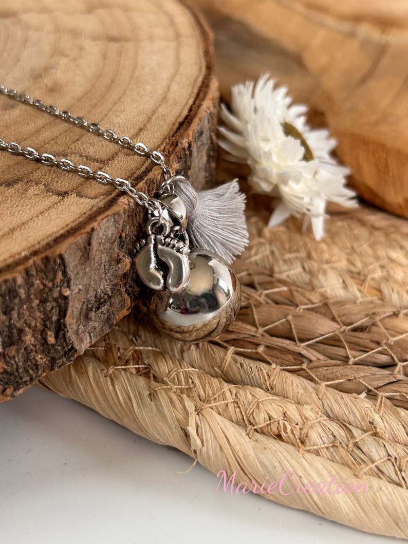 May include: Silver necklace with a silver charm of a baby's foot and a silver bell. The necklace has a gray tassel attached to it.