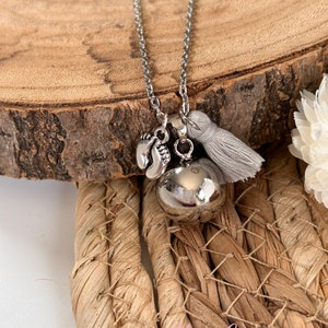 May include: Silver necklace with a silver bell charm, a small charm with two footprints, and a gray tassel. The necklace is on a wooden surface.