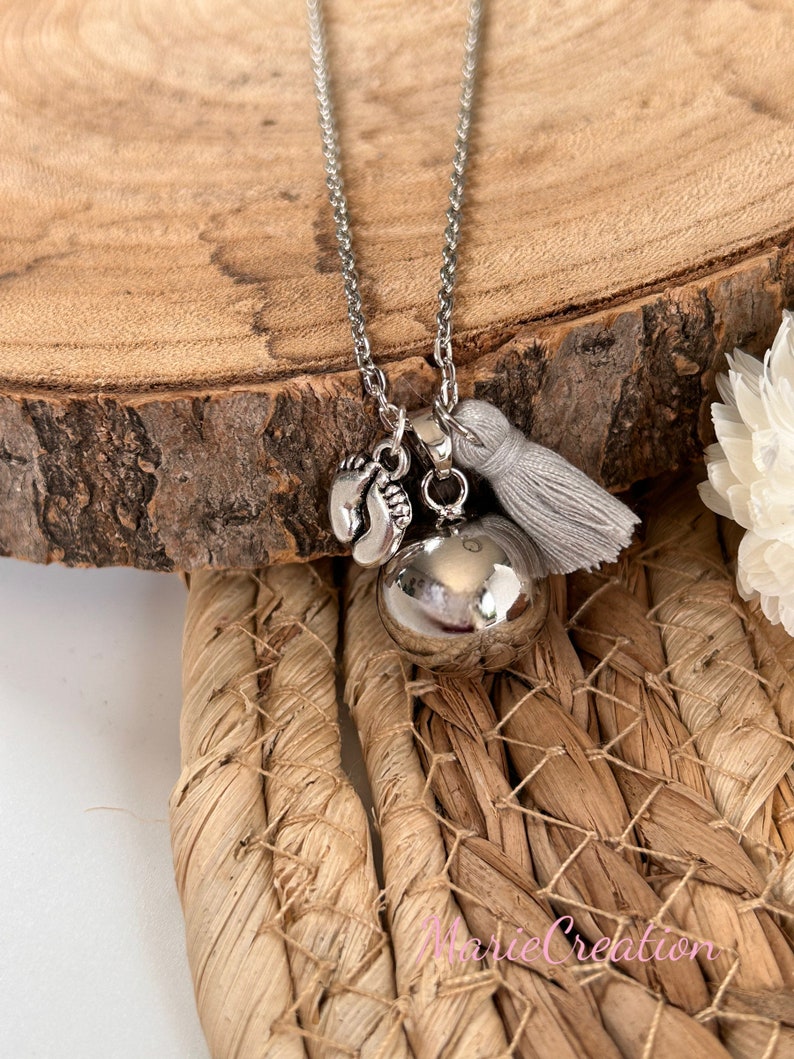 May include: Silver necklace with a silver bell charm, a small charm with two footprints, and a gray tassel. The necklace is on a wooden surface.
