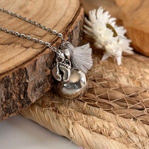 May include: A silver necklace with a charm of a small bell, a heart, and a tassel. The necklace is on a wooden surface with a white flower.