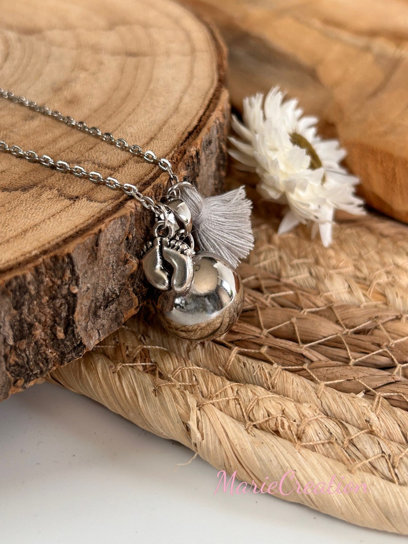 May include: A silver necklace with a charm of a small bell, a heart, and a tassel. The necklace is on a wooden surface with a white flower.
