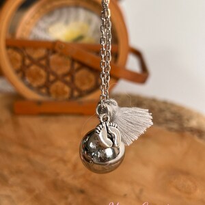 May include: Silver necklace with a silver charm in the shape of a baby's feet. The charm is attached to a silver ball that makes a sound when shaken. The necklace has a white tassel.