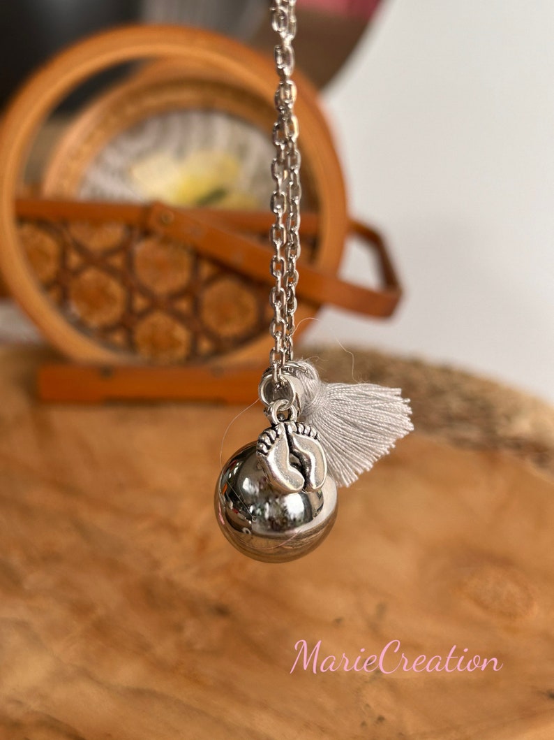 May include: Silver necklace with a silver charm in the shape of a baby's feet. The charm is attached to a silver ball that makes a sound when shaken. The necklace has a white tassel.