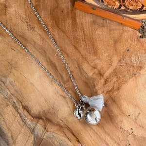 May include: A silver chain necklace with a silver charm, a silver baby foot charm, and a gray tassel. The necklace is on a wooden surface.