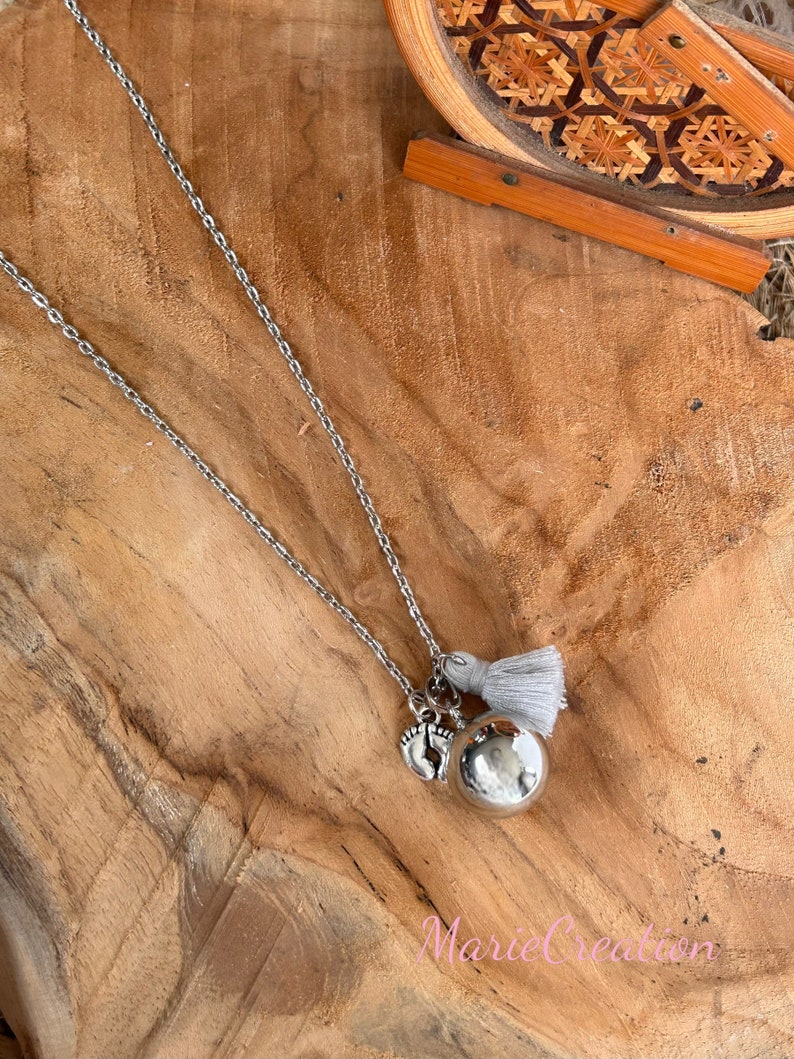 May include: A silver chain necklace with a silver charm, a silver baby foot charm, and a gray tassel. The necklace is on a wooden surface.