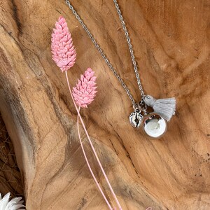 May include: Silver necklace with a silver charm of two tiny footprints and a silver bell with a gray tassel. The necklace is on a wooden surface with pink dried flowers.
