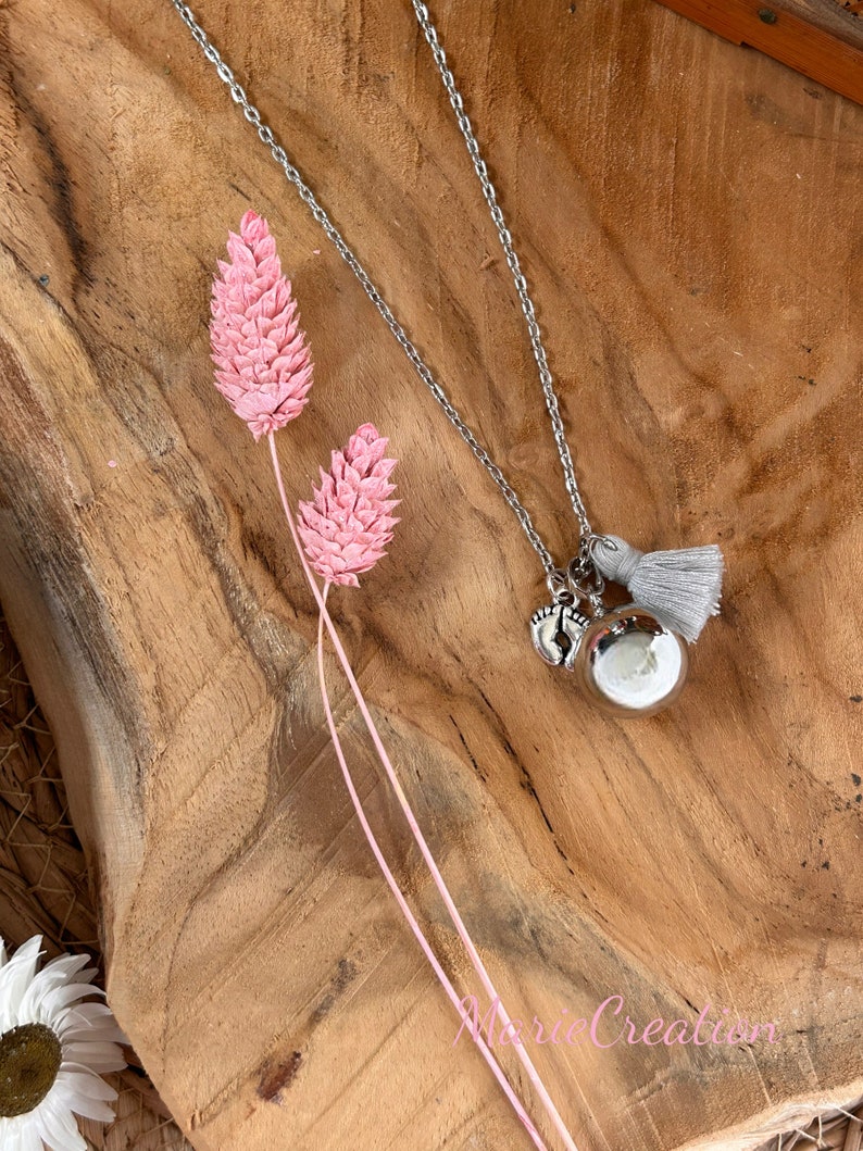 May include: Silver necklace with a silver charm of two tiny footprints and a silver bell with a gray tassel. The necklace is on a wooden surface with pink dried flowers.