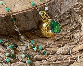 Könnte beinhalten: Eine goldfarbene Halskette mit einem Anhänger mit einem grünen tropfenförmigen Edelstein und einem runden, strukturierten Anhänger. Die Kette ist mit kleinen grünen, weißen und klaren Perlen verziert. Der Schmuck liegt auf einer Holzoberfläche und einer gewebten Matte.