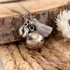 May include: Silver necklace with a silver baby feet charm, a silver bell charm, and a gray tassel charm. The necklace is on a wooden surface.