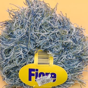 Könnte beinhalten: Ein Knäuel Fiore-Garn in Blau-, Weiß- und Grüntönen. Das Garn hat ein strukturiertes, flauschiges Aussehen. Ein gelbes Etikett mit dem Wort "Fiore" ist am Garn befestigt, zusammen mit einem Barcode und einem Preisschild.