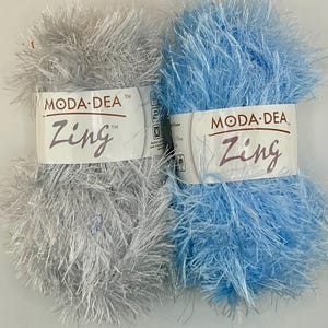 Pode incluir: Dois novelos de fio Moda Dea Zing. Um é de cor prata clara e o outro azul claro. O fio tem uma aparência difusa e texturizada. Os rótulos do fio dizem "Moda Dea" e "Zing".