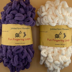 May include: Two skeins of Fun Fingering Loops yarn, one purple and one cream. Each has a yellow label with "Fun Fingering Loops" and "Jubilee Yarn Minnesota". The purple yarn is available in 43 colours, and the cream yarn in 48 colours. Ideal for knitting.