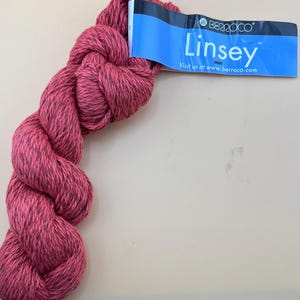 May include: A skein of twisted yarn in a deep rose-pink color with dark charcoal flecks throughout. Attached is a blue paper label with white text that reads Berroco Linsey and includes the website address www.berroco.com for product information.