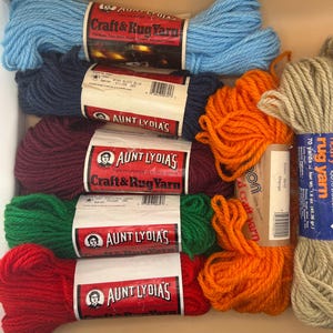 Puede incluir: Una colección de madejas de hilo en varios colores, incluyendo azul claro, azul oscuro, morado, verde, rojo, naranja y beige. Las etiquetas muestran las marcas Aunt Lydia's y Caron, con texto que indica 100% poliéster o Dacron.