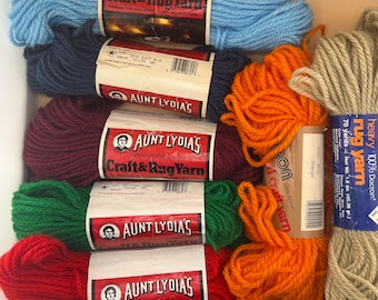 Könnte beinhalten: Eine Sammlung von Garnsträngen in verschiedenen Farben, darunter Hellblau, Dunkelblau, Lila, Grün, Rot, Orange und Beige. Die Etiketten zeigen die Markennamen Aunt Lydia's und Caron mit dem Hinweis auf 100% Polyester oder Dacron.