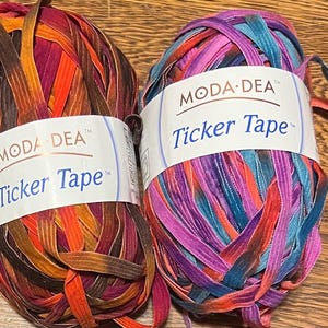 Pode incluir: Duas bobinas de fita Ticker Tape da Moda Dea. Uma bobina apresenta tons de laranja, vermelho e castanho. A outra bobina tem tons de roxo, azul e rosa. Cada bobina tem uma etiqueta branca com o logótipo Moda Dea e as palavras "Ticker Tape".