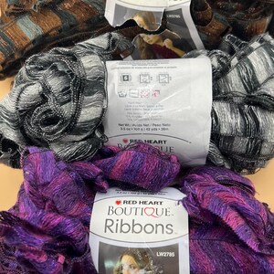 Może przedstawiać: Trzy motki przędzy Red Heart Boutique Ribbons w różnych kolorach: brązowym i szarym, czarnym i srebrnym oraz fioletowym i różowym. Każdy motek ma etykietę z nazwą marki i informacjami o produkcie.