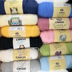 May include: A collection of ten Caron Simply Soft yarn skeins in various colours including navy, cream, yellow, black, white, grey, berry blue, pink, light pink, lime green, and light blue, each with a paper label showing 170g or 141.7g weight.