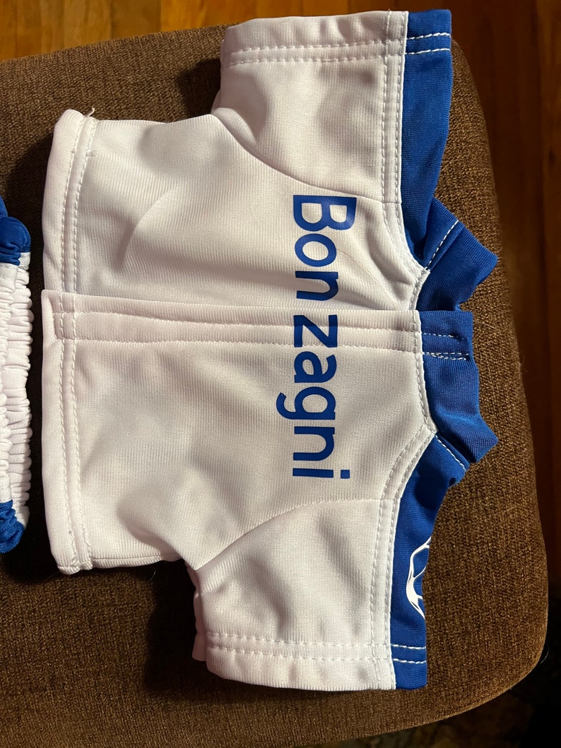 Puede incluir: Camiseta de osito de peluche blanca y azul con el texto "Bon Zagni!" impreso en la parte delantera.