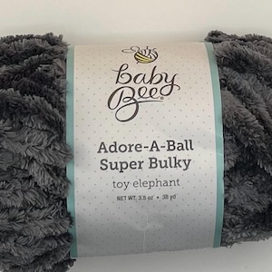 May include: A skein of Baby Bee Adore-A-Ball Super Bulky yarn in a soft, gray color. The label reads "toy elephant" and the net weight is 3.5 oz. The yarn is fluffy and appears ideal for crafting.