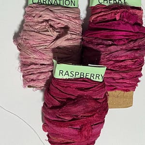 K&ouml;nnte beinhalten: Drei Spulen mit ausgefranstem Seidenband in Rosa- und Rott&ouml;nen. Die obere linke Spule ist mit Pink Carnation beschriftet, die obere rechte mit Cherry und die untere mittlere Spule mit Raspberry. Jede Spule weist raue Kanten und Textur auf.