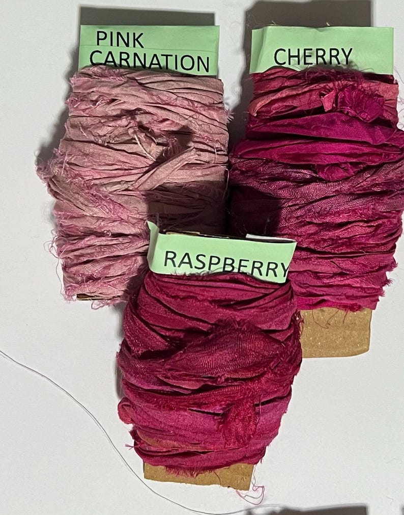 K&ouml;nnte beinhalten: Drei Spulen mit ausgefranstem Seidenband in Rosa- und Rott&ouml;nen. Die obere linke Spule ist mit Pink Carnation beschriftet, die obere rechte mit Cherry und die untere mittlere Spule mit Raspberry. Jede Spule weist raue Kanten und Textur auf.