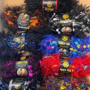 Puede incluir: Una colección de ovillos de lana Lion Brand Fancy Fur y Festive Fur en varios colores, incluyendo negro, naranja, azul, rojo y marrón. Cada ovillo presenta una etiqueta de papel negro con el nombre del producto y el logotipo.