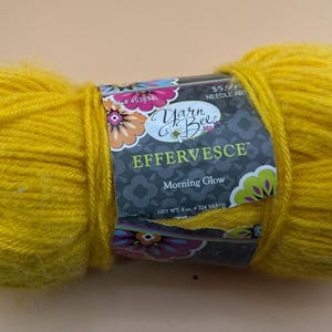 Könnte beinhalten: Ein leuchtend gelber Knäuel Yarn Bee Effervescence-Garn in der Farbe Morning Glow. Das Etikett zeigt florale Designs und den Text "Effervescence" und "Morning Glow". Das Garn wiegt 113g und hat eine Lauflänge von 205 Metern.