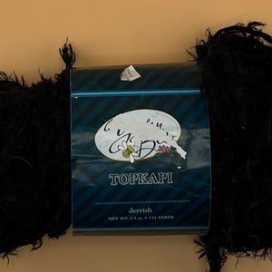 May include: A ball of black yarn with a Topkapi yarn package in the middle. The Topkapi package is dark blue with white text and a floral design. The yarn is fluffy and looks to be made of a soft material.